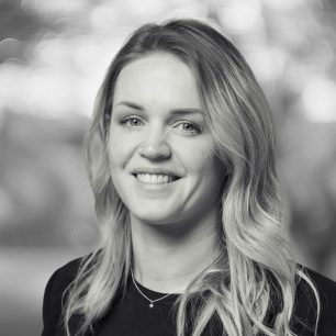 A woman with long, wavy hair smiles in a black and white portrait, with a blurred background.