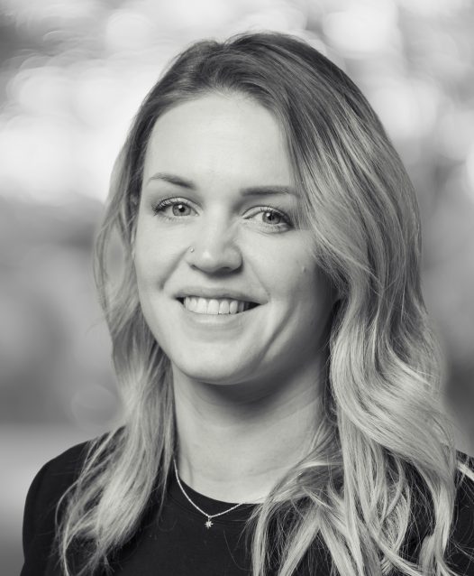 A woman with long, wavy hair smiles in a black and white portrait, with a blurred background.