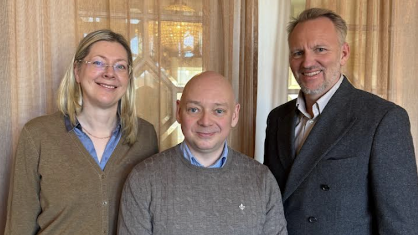 Three people standing together indoors, smiling at the camera.