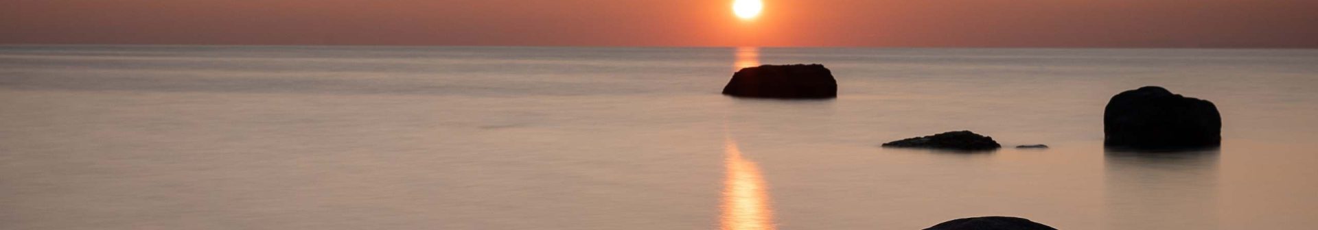 The sun sets over a calm sea with large rocks in the foreground, casting a warm orange glow across the sky and water.