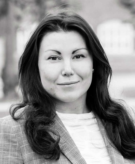 A woman with long hair wearing a plaid blazer and a white top, standing outdoors with blurred buildings in the background.