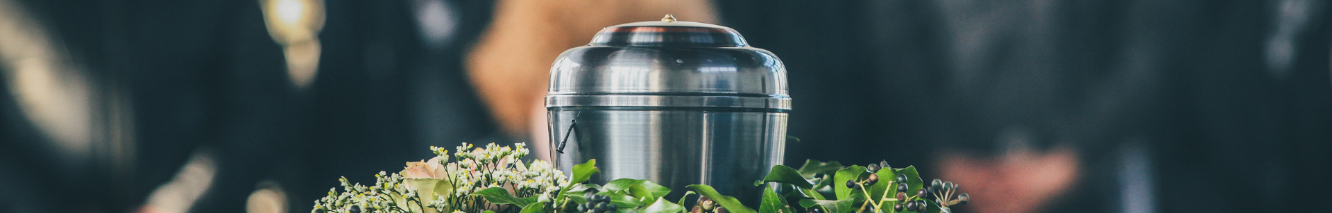 A silver urn is placed on a surface surrounded by green leaves and small flowers, with blurred dark figures in the background.