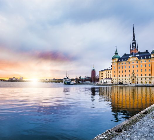 A waterfront scene at sunset with a large historical building featuring green domes and a tall spire reflecting on calm water, with a cobblestone path in the foreground.