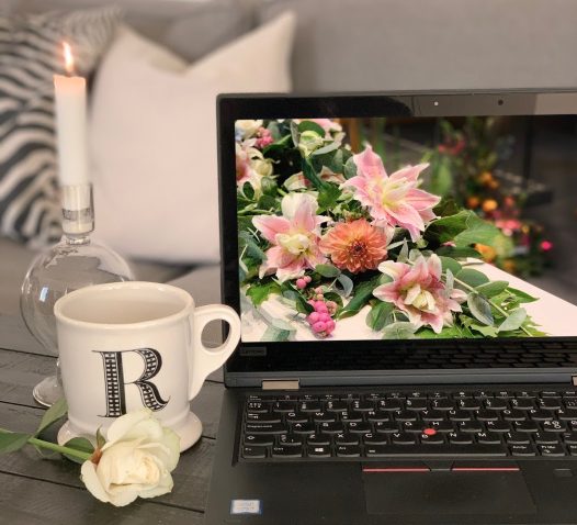A laptop displaying an image of pink flowers is on a wooden table, next to a lit candle and a mug with the letter "R," adorned with a white rose.