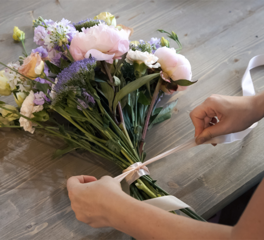 Hands tying a white ribbon around a bouquet of mixed flowers, including pink and purple blooms, on a wooden table.