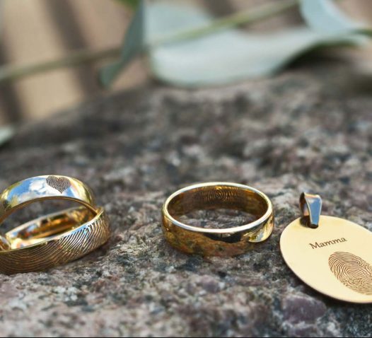 Two gold rings and an oval pendant with fingerprint engravings rest on a rock. The pendant is inscribed with the word "Mamma" and a fingerprint design.
