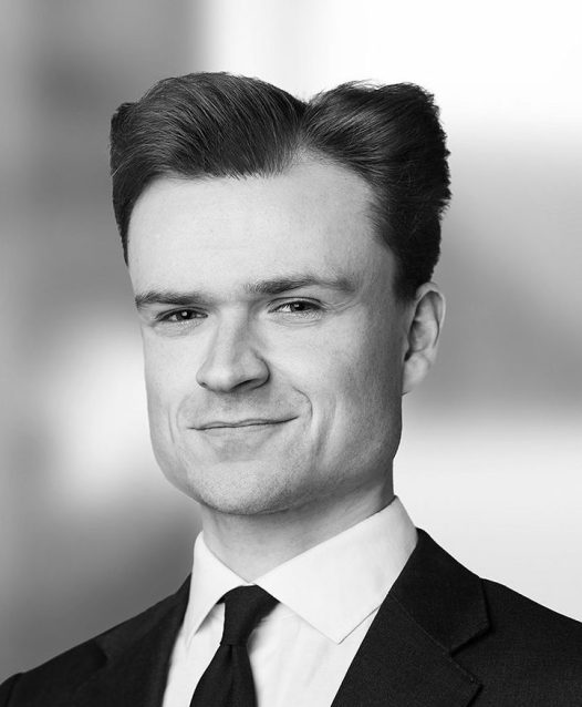 A person in a suit and tie smiles slightly, with a blurred background.